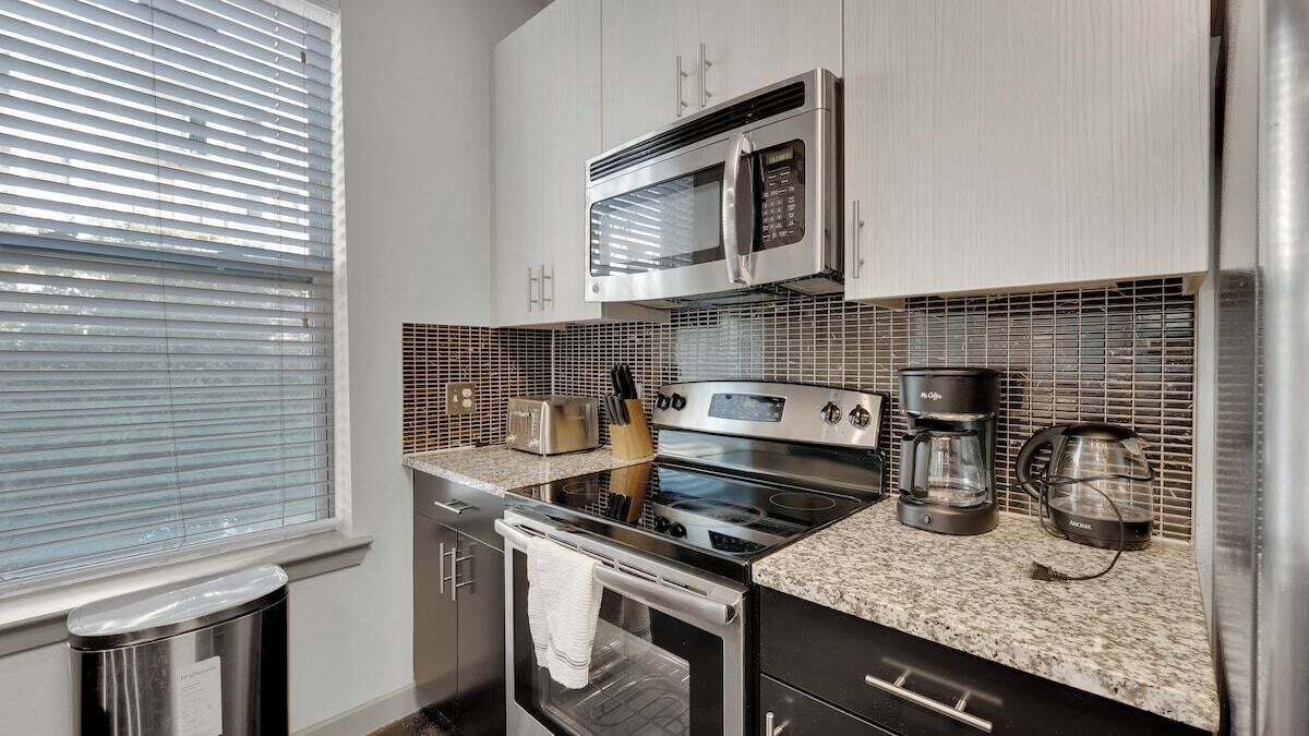 A modern kitchen with stainless steel appliances, a granite countertop, and a window with blinds.