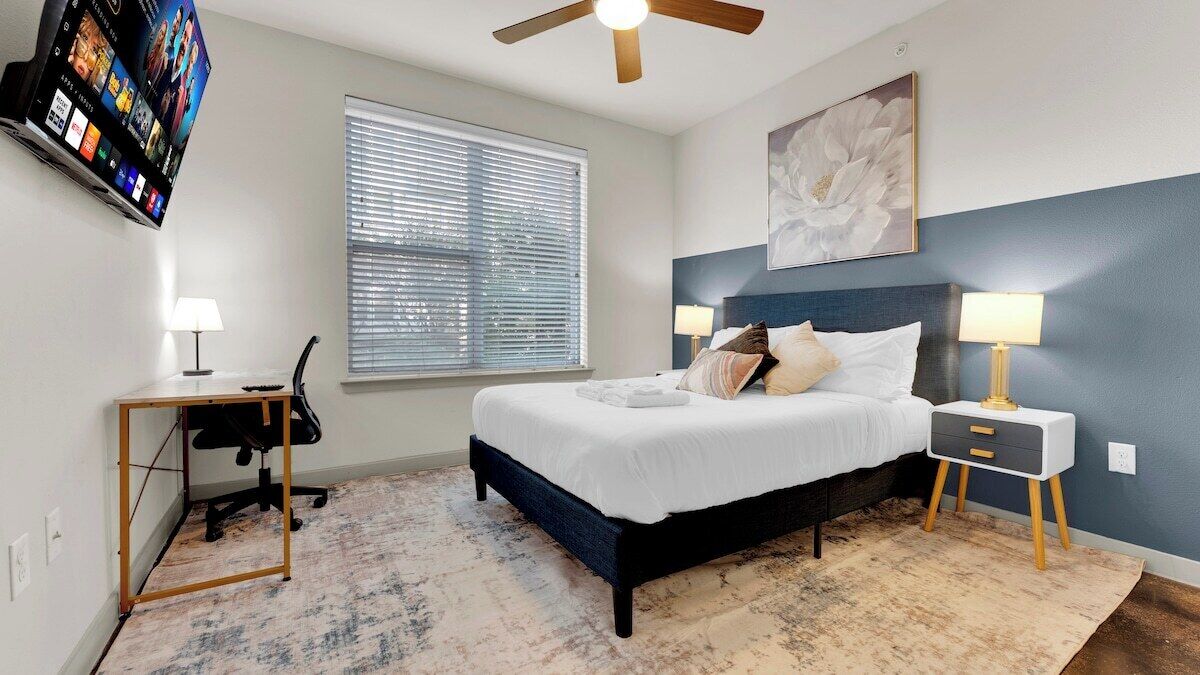 A bedroom with a bed, bedside tables, a desk, a chair, a ceiling fan, a window with blinds, and a wall-mounted TV.