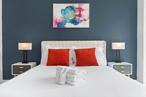 A hotel room with a bed, two nightstands, a white headboard, red pillows, a white blanket, and a colorful abstract painting on the wall. - Contemporary 1BR | Sleeps 2 | Northern Liberties (Philadelphia)