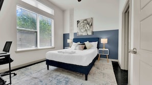 A bedroom with a bed, bedside tables, a desk, and a window with blinds. - Luxury 2BR Uptown | Sleeps 12 | Parking (Dallas)