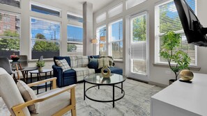 A modern living room with a sectional sofa, a glass coffee table, and a view of the outdoors through large windows. - Luxury 2BR Uptown | Sleeps 12 | Parking (Dallas)