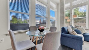 A modern living room with a glass table, grey chairs, a blue sofa with cushions, and large windows with blinds. - Luxury 2BR Uptown | Sleeps 12 | Parking (Dallas)