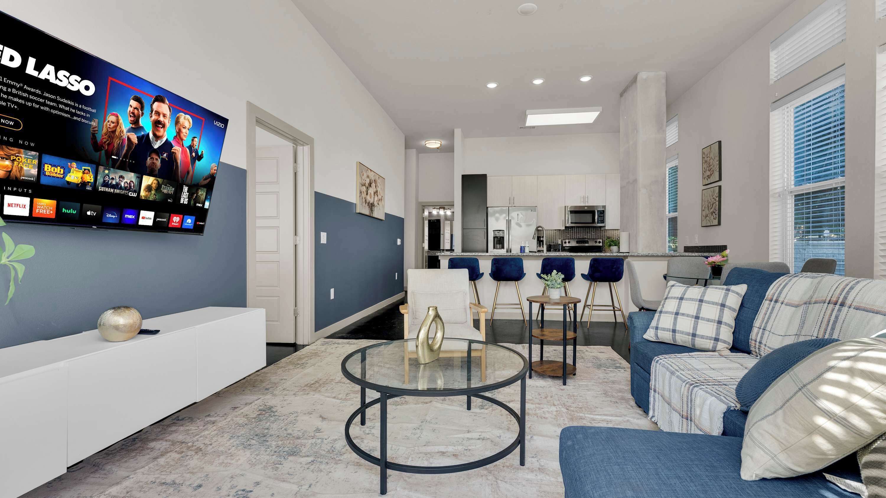 A modern living room with a flat-screen TV, a sofa, a coffee table, and a kitchen in the background.