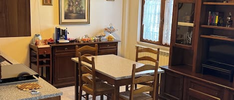 Una cocina con armarios de madera, una mesa de comedor con sillas, un ventanal con cortinas de encaje y una estantería.