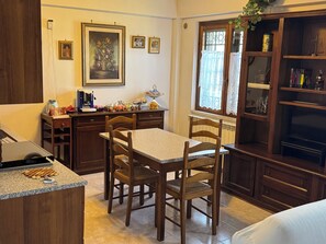 A kitchen with wooden cabinets, a dining table with chairs, a window with lace curtains, and a bookshelf.