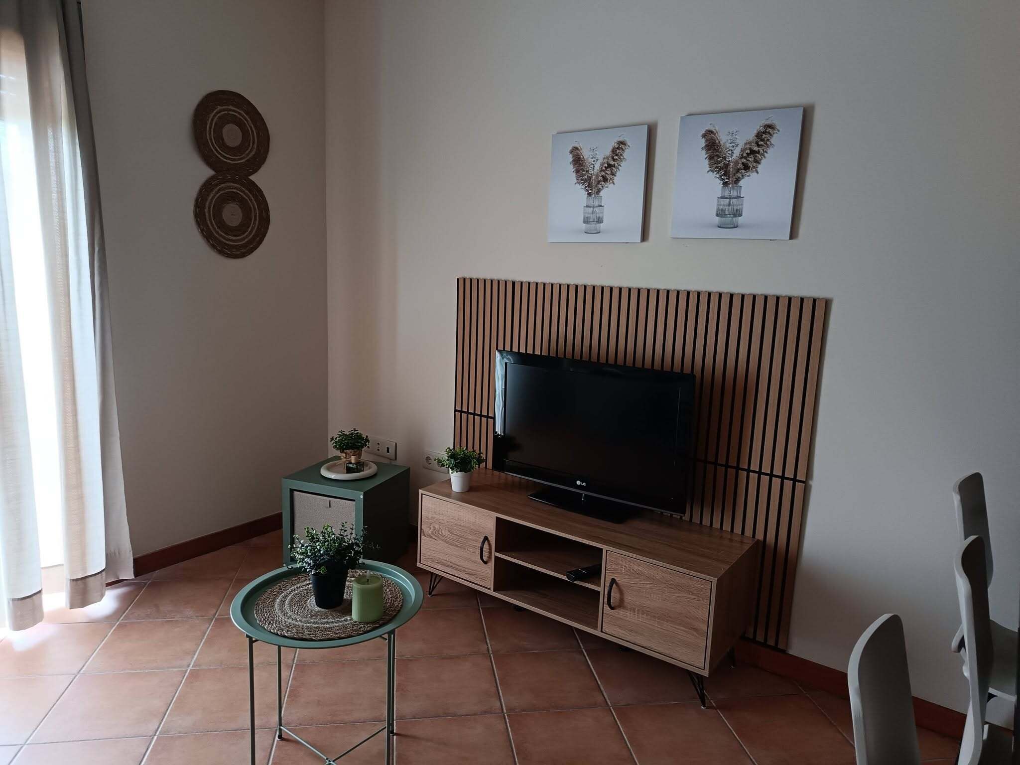 Un saló amb un televisor de pantalla plana sobre un moble de televisió de fusta, una taula petita amb una planta i dues imatges penjades a la paret.