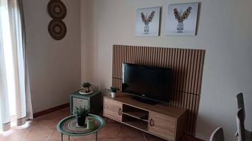 Un saló amb un televisor de pantalla plana sobre un moble de televisió de fusta, una taula petita amb una planta i dues imatges penjades a la paret.