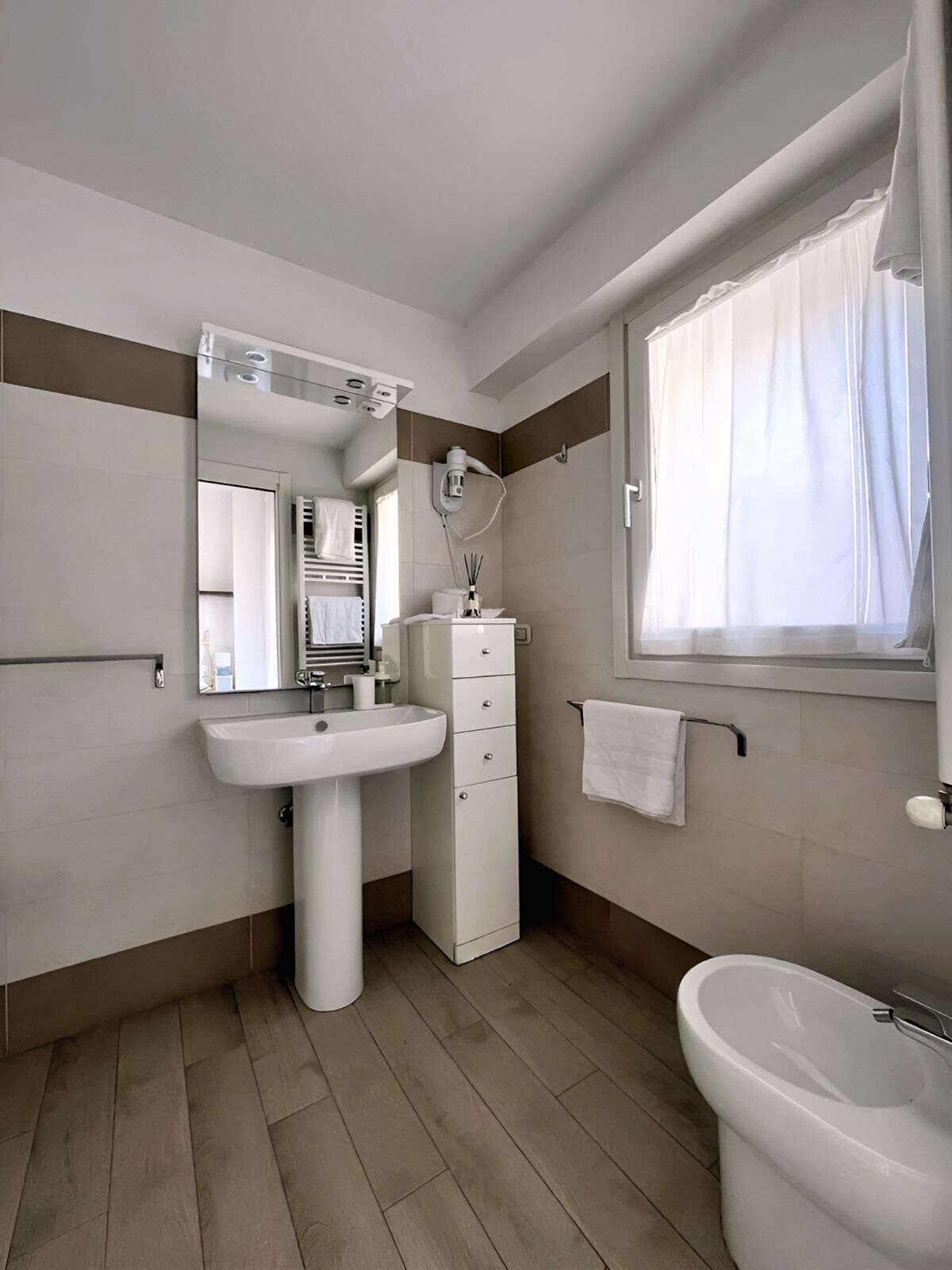 Un bagno moderno con un lavabo su piedistallo, uno specchio, un porta asciugamani e una finestra con tende.