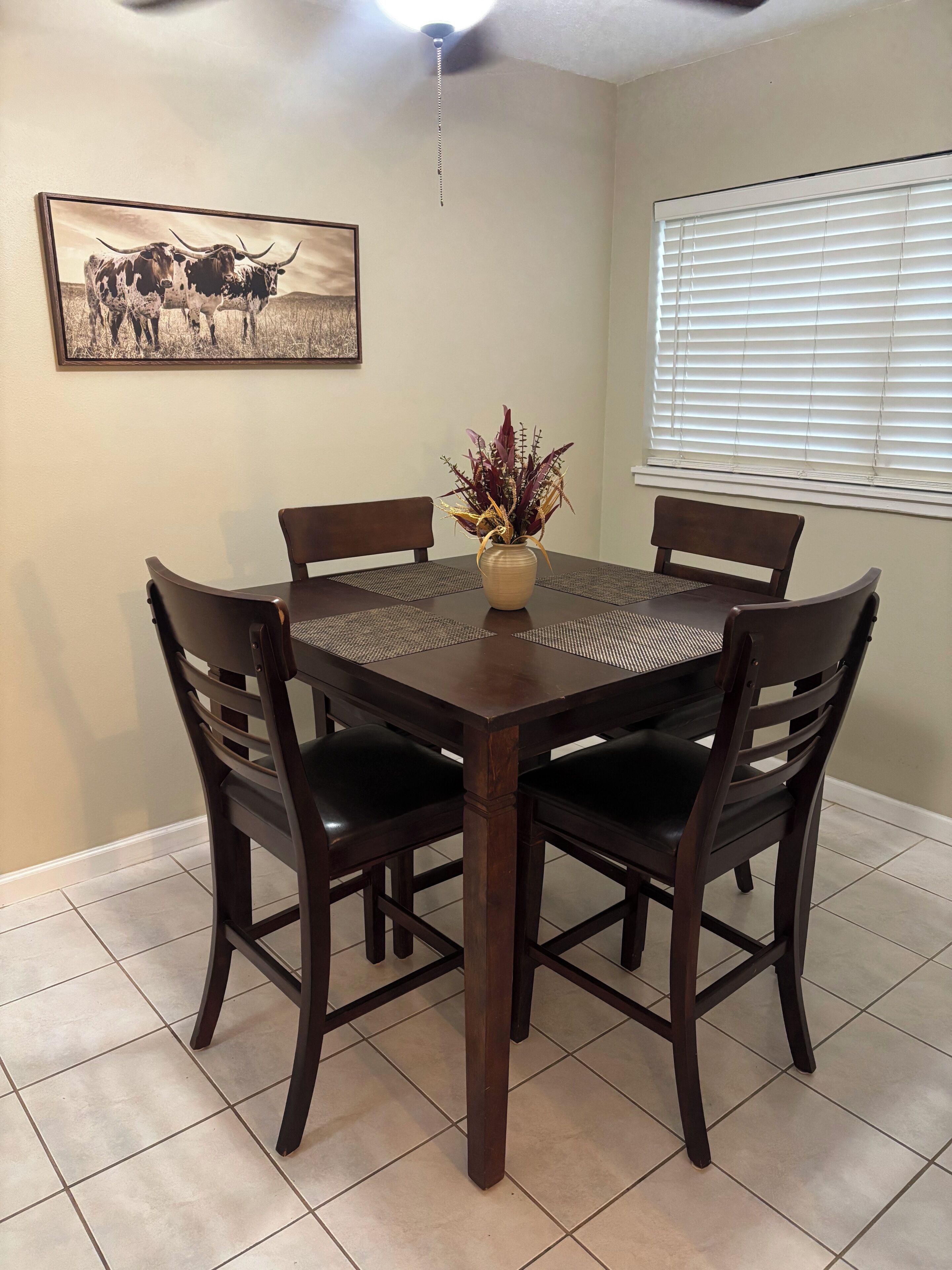 A dining area with a round table, six chairs, a vase with dried plants, a ceiling fan, a window with blinds, and a framed picture on the wall.