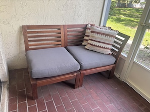 A wooden bench with cushions and a patterned throw pillow, positioned on a brick floor with a view of a grassy area through a window.