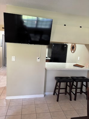 A modern kitchen with a flat-screen TV mounted above the counter, a refrigerator, and two bar stools.