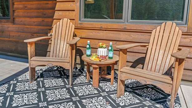 Dos sillas Adirondack de madera, una mesita redonda con botellas y fruta, y una alfombra estampada en un patio.