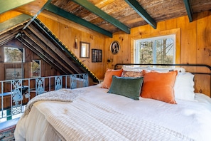 Un dormitorio acogedor con techo inclinado, una cama con muchos cojines, una ventana con vista a árboles y un pequeño balcón con barandilla.