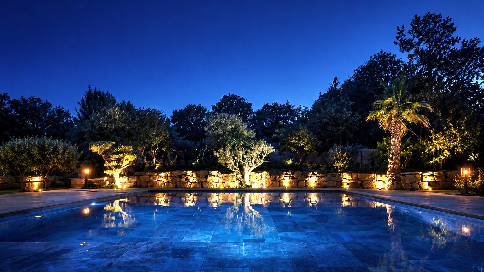 A nighttime swimming pool with stone walls and trees, illuminated by ambient lighting.