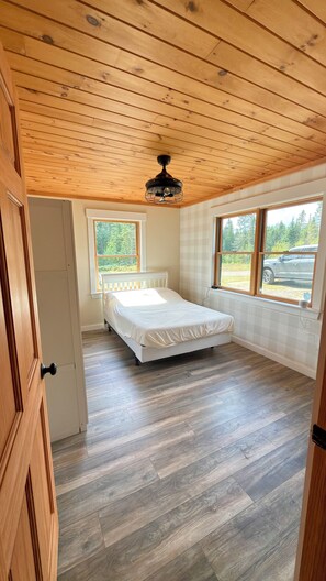 A room with a bed, wooden flooring, a ceiling fan, and windows with a view of the outdoors. - Charming 3-bedroom house in serene Eustis perfect for relaxing stays! (Eustis)