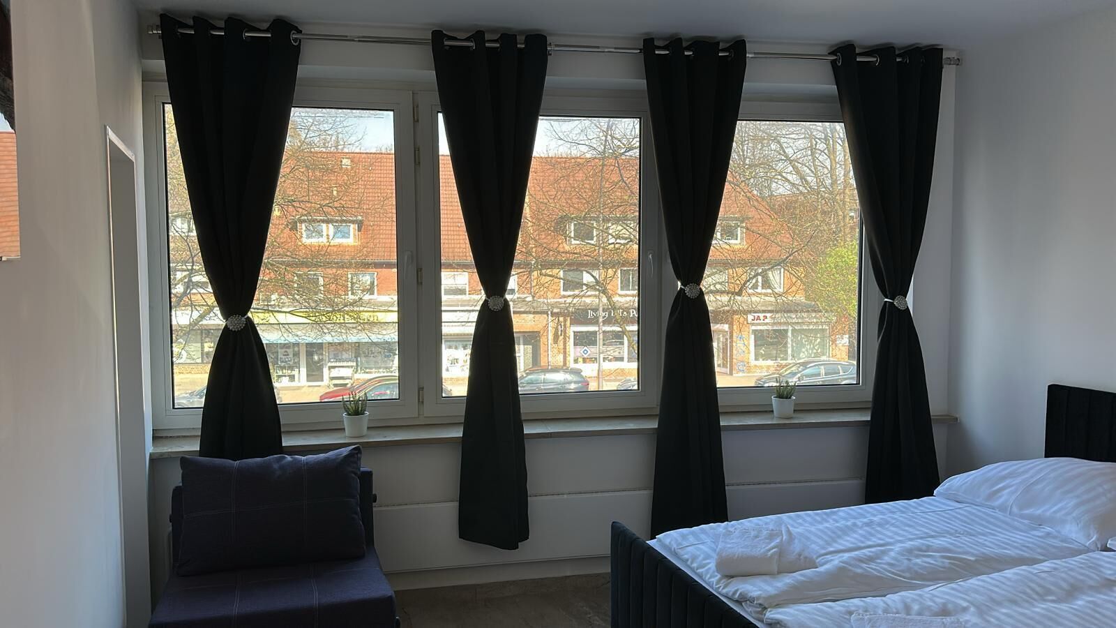 Ein Schlafzimmer mit einem Bett, einem Fenster mit Vorhängen, einem kleinen Tisch mit Pflanze und Blick auf Gebäude.