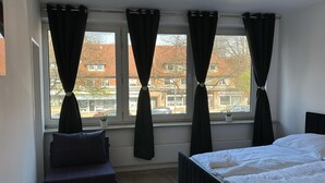 Een slaapkamer met een bed, een raam met gordijnen, een tafeltje met een plant en uitzicht op gebouwen.