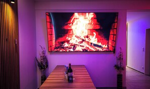Une pièce avec un grand téléviseur à écran plat diffusant une image de feu, une table en bois avec une bouteille et des verres, et deux plantes en pot.