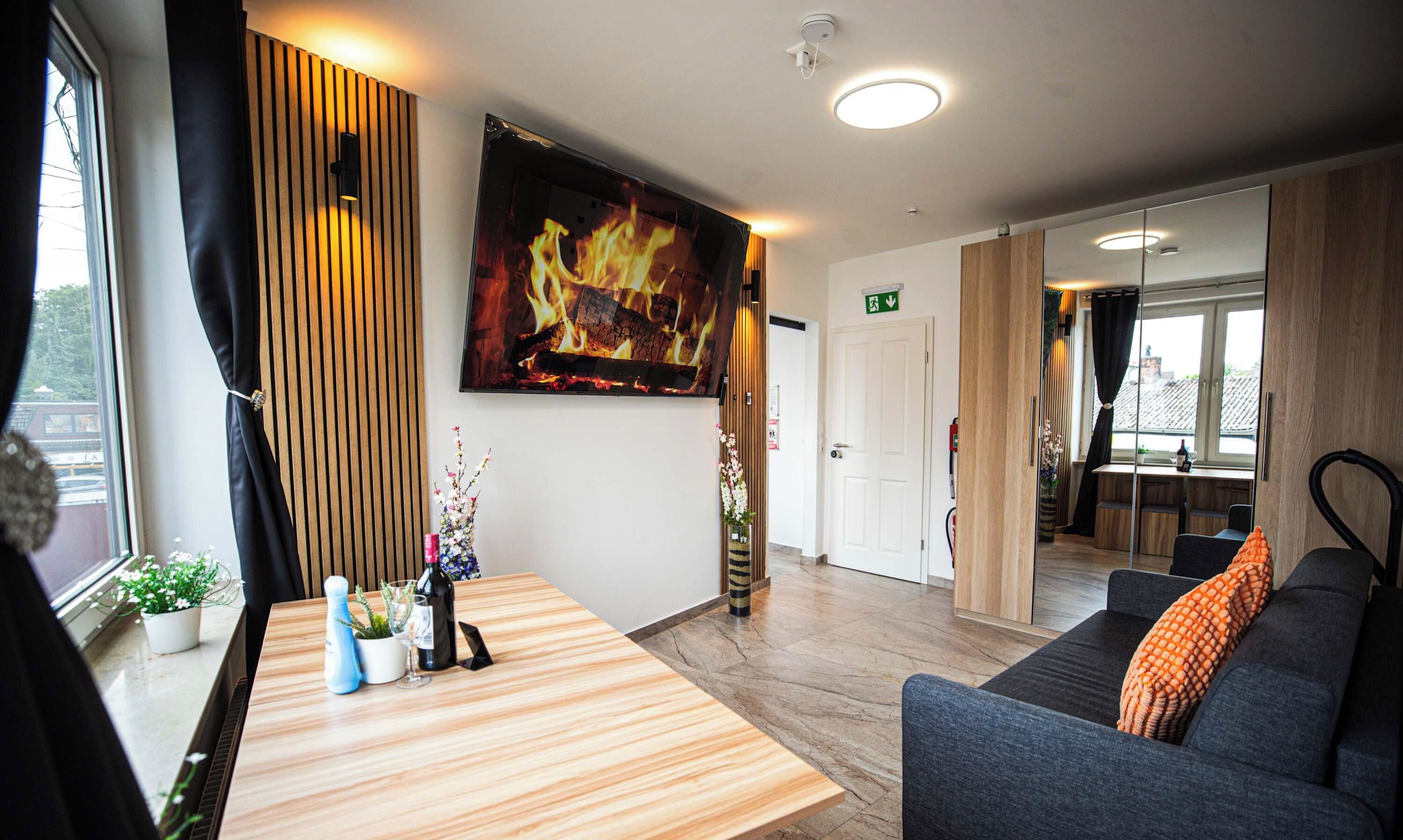 Een moderne hotellobby met een houten receptiebalie, een zithoek met een bank, een grote flatscreen-tv waarop een haard te zien is, en uitzicht naar buiten door grote ramen.