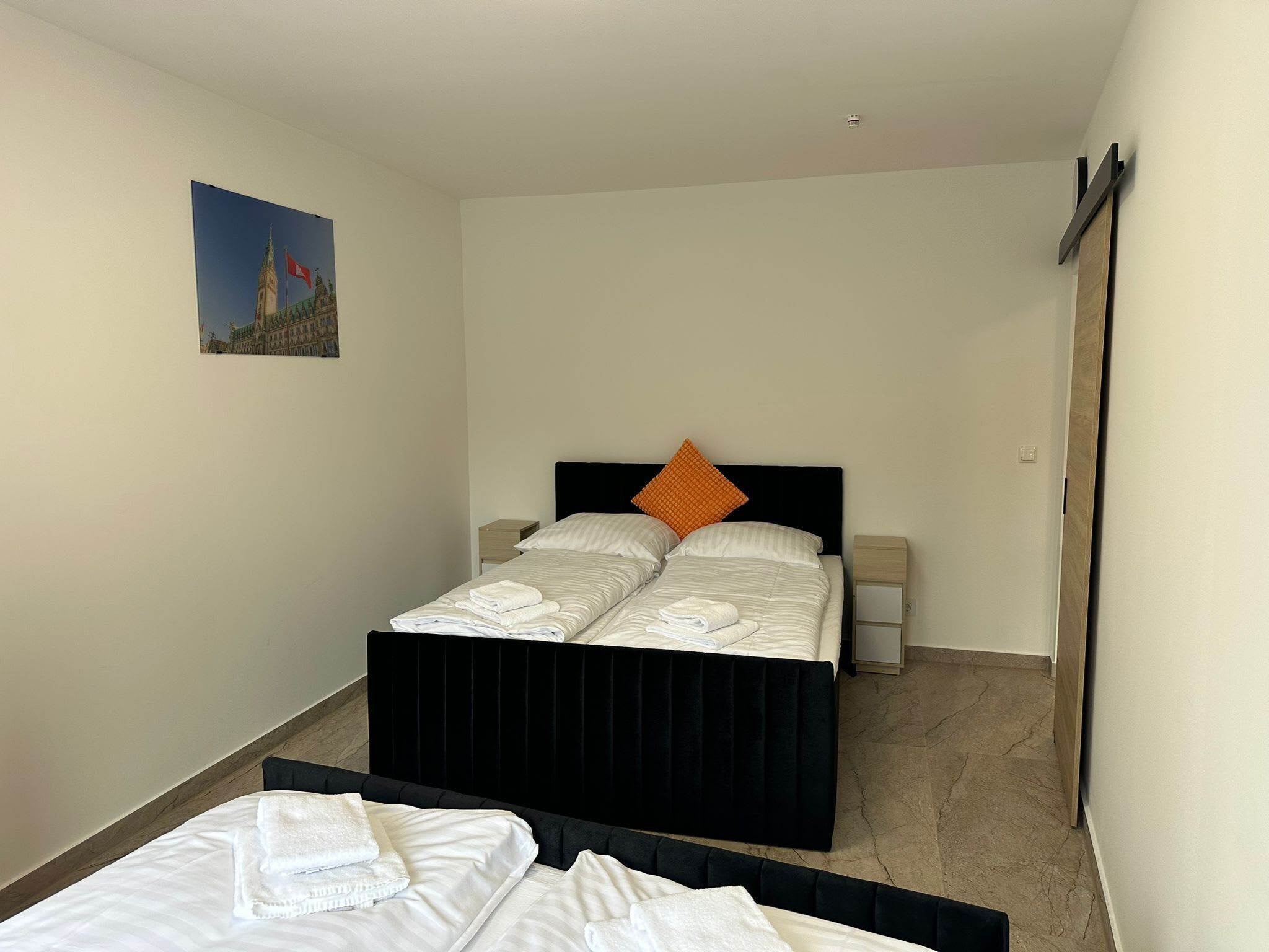 Ein Hotelzimmer mit Doppelbett, zwei Nachttischen, einem Beistelltisch und einem Bild von einem Gebäude an der Wand.