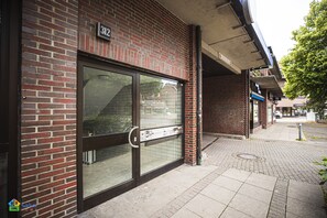 Un bâtiment en briques avec une plaque de numéro de maison, une porte en verre dotée d’une poignée en métal et un trottoir pavé.