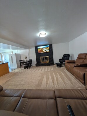 A living room with a fireplace, a television mounted above it, a brown leather sofa, and a dining area with chairs and a table.