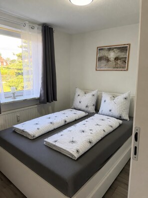 Uma cama de casal com travesseiro listrado e uma cama simples com travesseiro liso em um quarto com janela e pintura na parede.