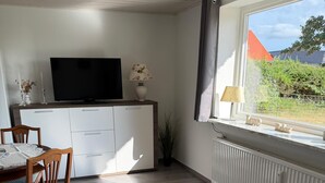 Ein modernes Wohnzimmer mit Flachbildfernseher, einem Esstisch mit Stühlen, einem weißen Schrank und einem Fenster mit Blick auf Grünflächen.
