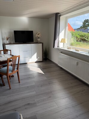 A modern living room with a flat-screen TV, a dining table with chairs, a white cabinet, and a window with a view of greenery. - "Reethus – A Little Getaway" Vacation Rental in a Quiet Setting, Near the North Sea and the Islands (Bosbüll)