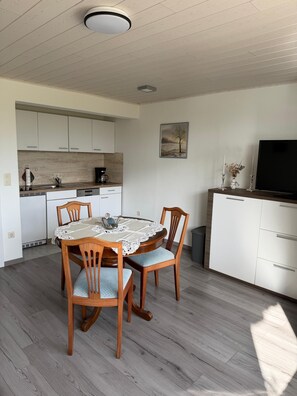 A compact kitchen with white cabinets, a dining table with chairs, and a flat-screen TV on a stand. - "Reethus – A Little Getaway" Vacation Rental in a Quiet Setting, Near the North Sea and the Islands (Bosbüll)