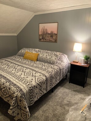Een slaapkamer met een bed, een nachtkastje, een lamp, een schilderij van een cactus en een kussen met tekst.