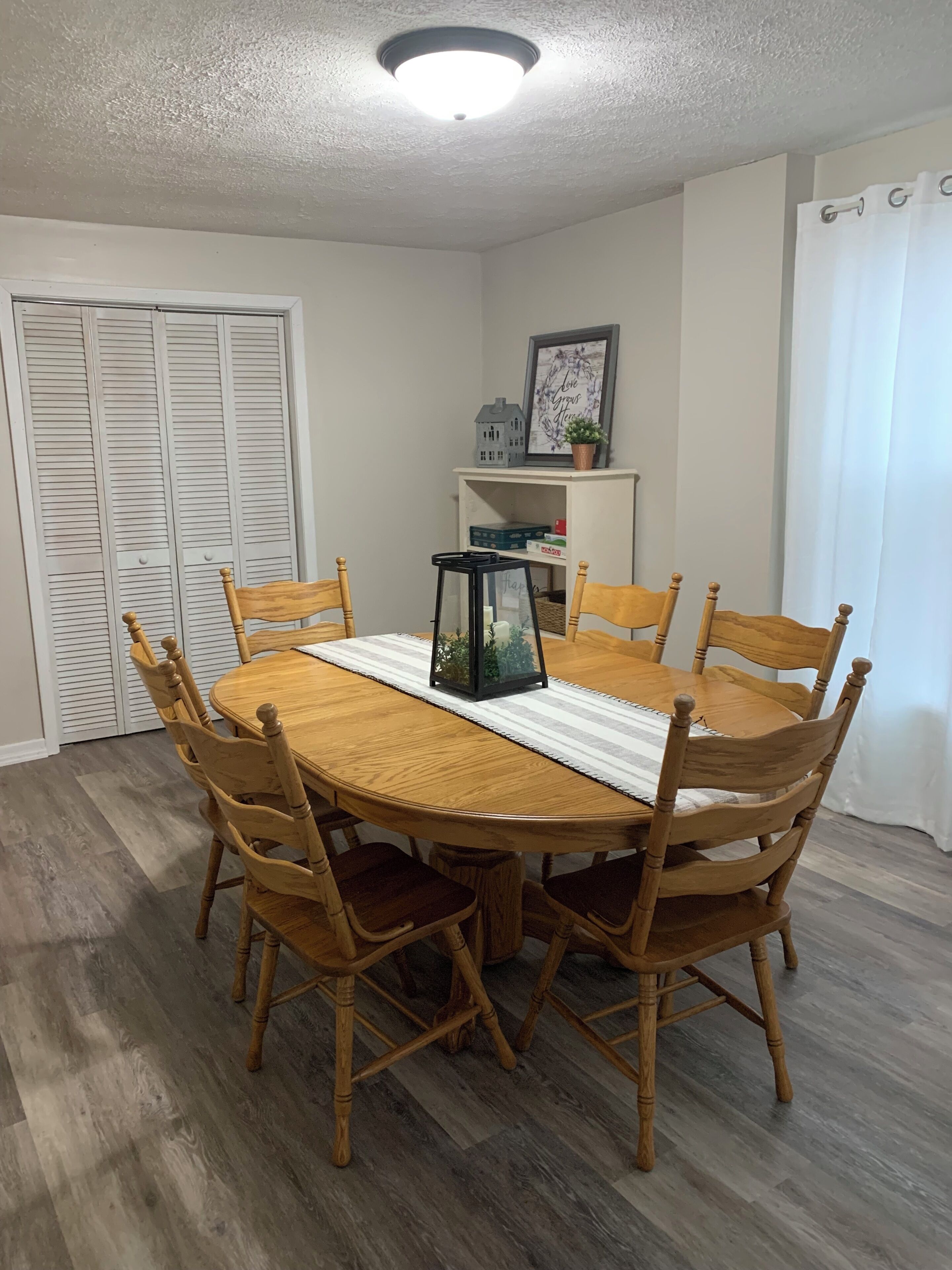 A dining area with a round wooden table surrounded by six wooden chairs, a black lantern, and a shelf with decorative items.
