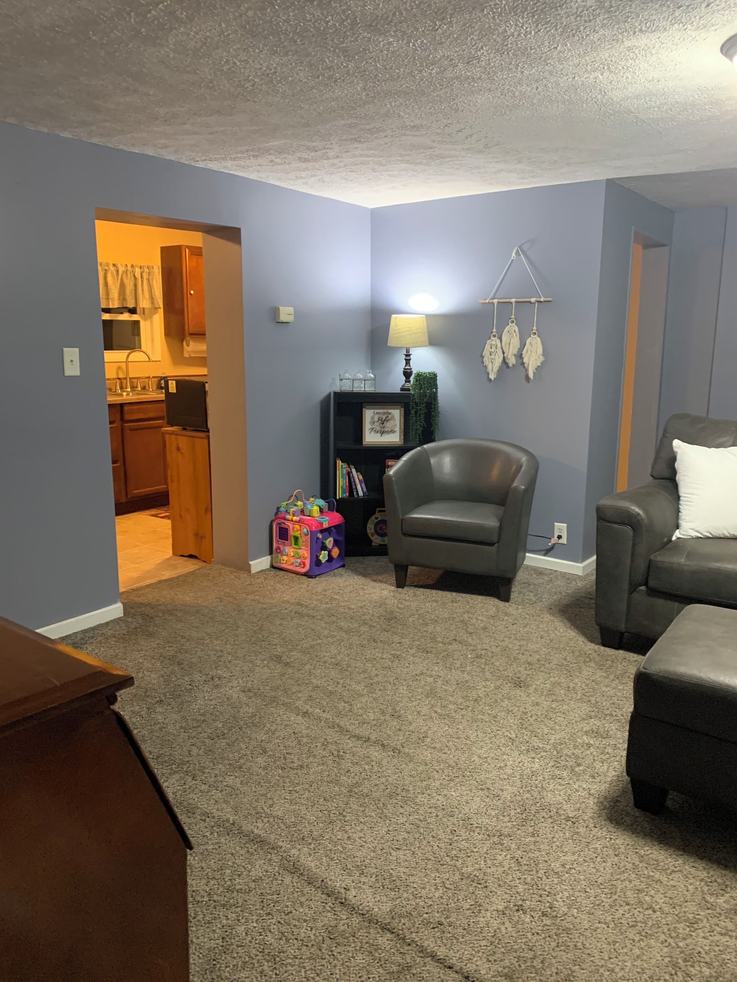 A living room with a grey armchair, a sofa, a small table, and a bookshelf with decorative items.