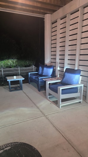 Eine Außenpatio mit einer Sitzgruppe aus einer Couch und zwei Stühlen, einem kleinen Tisch und einem Metallzaun im Hintergrund.