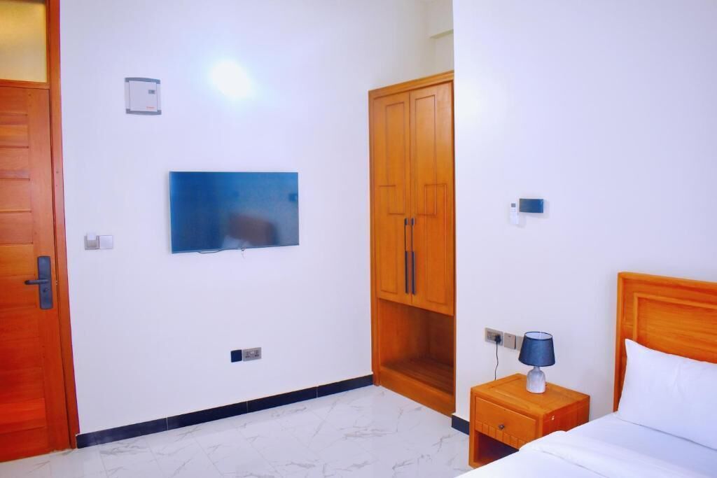 Habitación de hotel con cama, mesita de noche con lámpara, armario, televisor montado en la pared y una puerta.