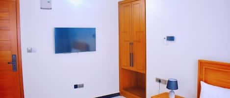 Habitación de hotel con cama, mesita de noche con lámpara, armario, televisor montado en la pared y una puerta.