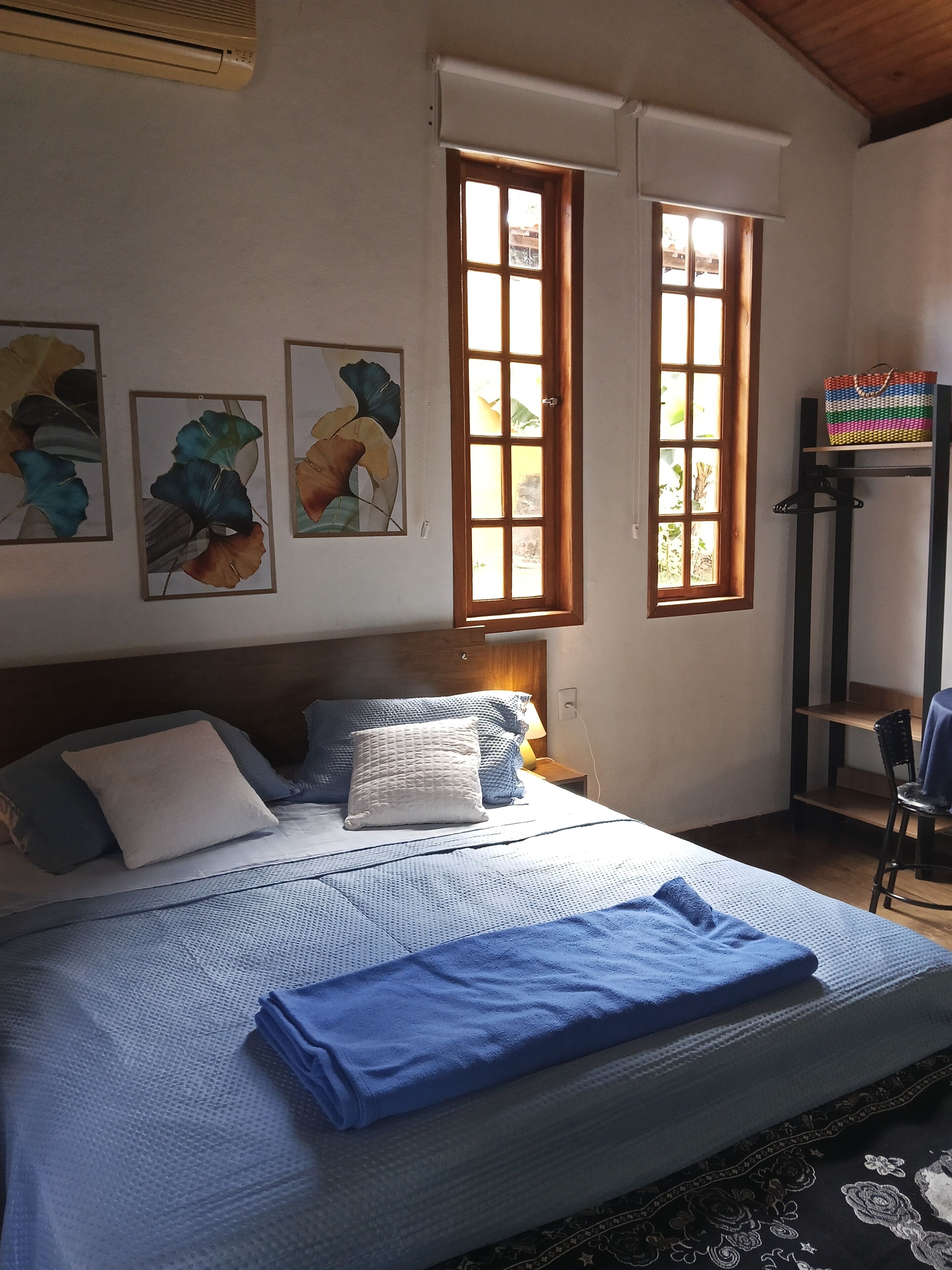 A neatly made bed with blue bedding, a headboard, and a nightstand with a lamp. There are two windows with wooden frames and a shelf with a colorful bag.