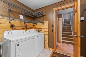A laundry area with two white front-loading washers and dryers, wooden paneling, metal shelving, and a wooden door leading to a staircase.