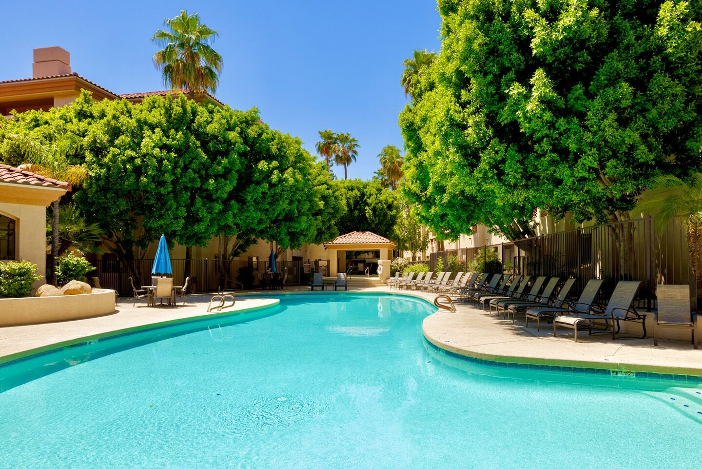 A swimming pool with lounge chairs and umbrellas, surrounded by trees and buildings.