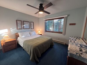 Ein Schlafzimmer mit einem Bett, einem Nachttisch, einer Lampe, einem Deckenventilator, einem Fenster mit Blick auf Bäume und einer Decke auf dem Boden.
