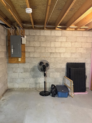 Un sous-sol avec des poutres en bois apparentes, un ventilateur sur pied, un portail en métal noir et une boîte de rangement bleue.