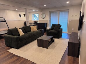 Un salon avec un canapé vert foncé, un fauteuil plus petit, une table basse et une grande fenêtre avec des stores.