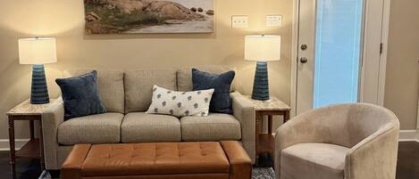 A living room with a beige sofa, two side tables with lamps, a brown ottoman, a patterned rug, and a door with a window.