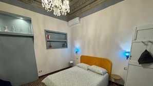 A bedroom with a patterned carpet, a bed with a patterned bedspread, a chandelier, a wardrobe, and a small table with a lamp.
