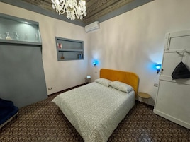 Un dormitorio con alfombra estampada, cama con colcha estampada, candelabro, armario y una mesita de noche con lámpara.