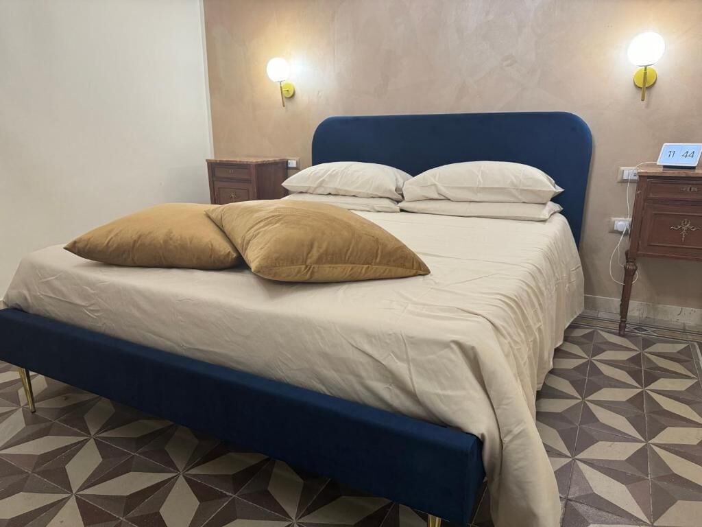 Uma cama com cabeceira azul, duas almofadas e uma mesa de cabeceira com abajur.