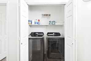 A laundry room with two front-loading washers and dryers, a shelf with cleaning supplies, and a decorative sign.