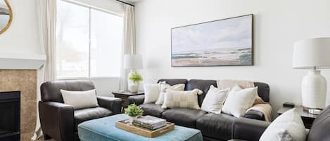 A living room with a fireplace, a large window with curtains, a sofa set, a coffee table, and a painting on the wall.