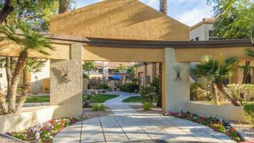 A covered outdoor area with a pathway leading to a building, surrounded by palm trees and a landscaped garden.