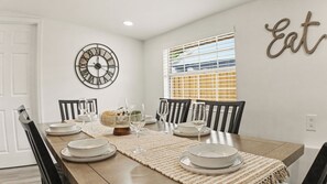A dining room with a wooden table set for four, surrounded by chairs, with a decorative wall clock and a window with blinds. - Tampa Family Home with Putting Green + Fenced Yard (Tampa)